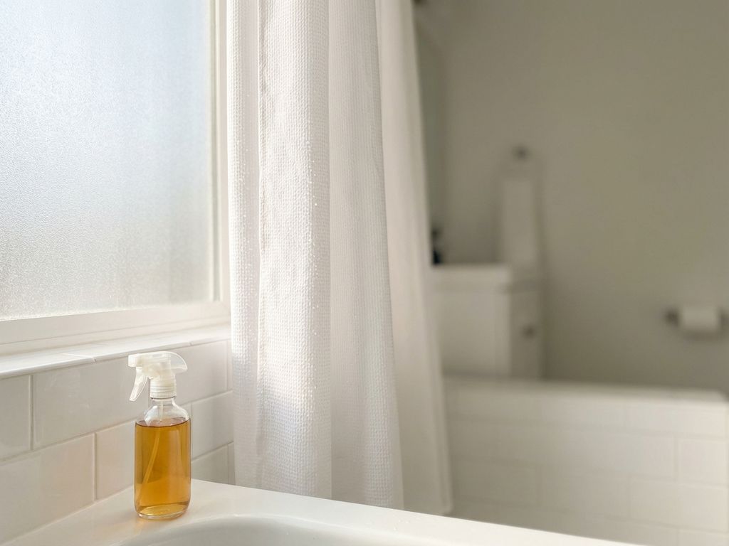 Glazen spuitfles met azijn op badrand, witte douchegordijn met waterdruppels op achtergrond in moderne badkamer